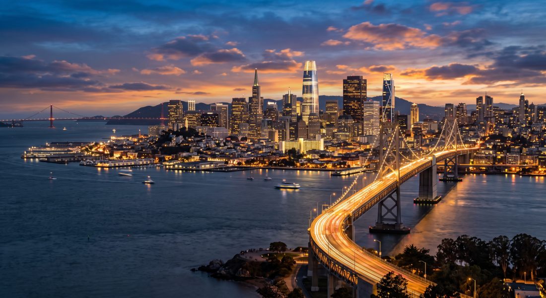 San Francisco skyline and the Bay Bridge at dusk, with Salesforce Tower and the Transamerica Pyramid against a sunset sky